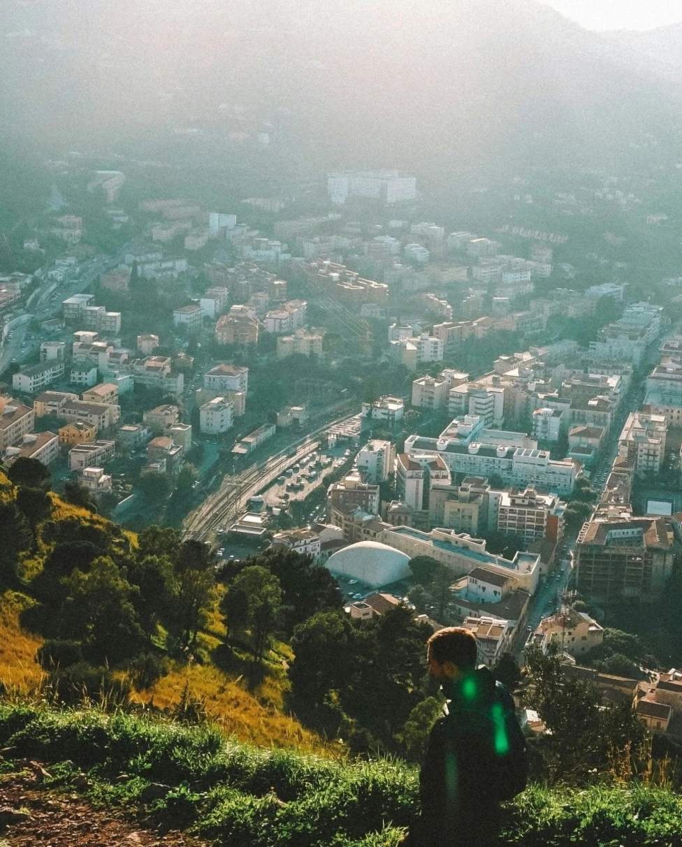 A person hiking in Sicily on a grassy hill with the city, ocean and mountains in the background on a sunny day.