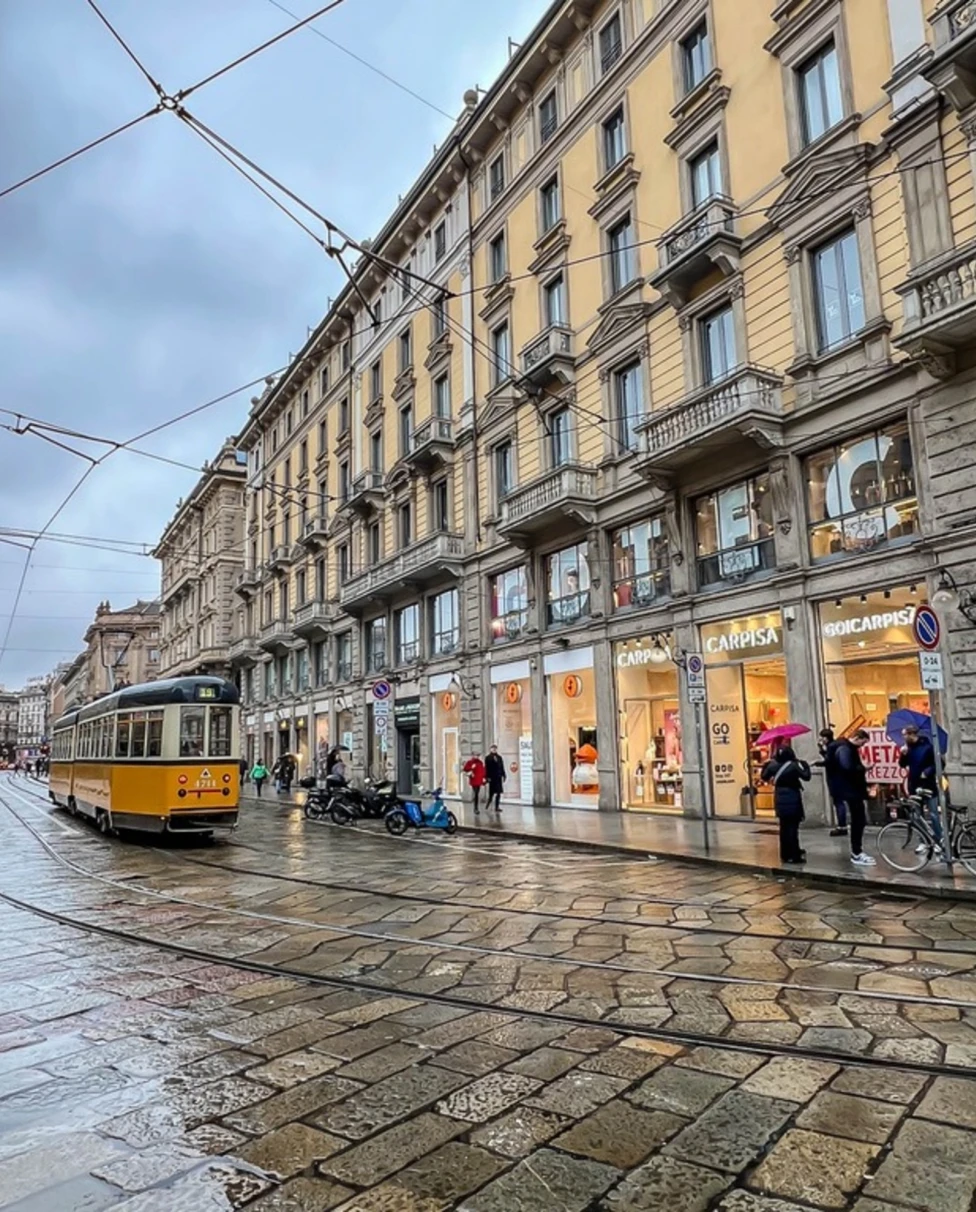 A wet urban street scene with a yellow tram, pedestrians, and classic architecture under an overcast sky