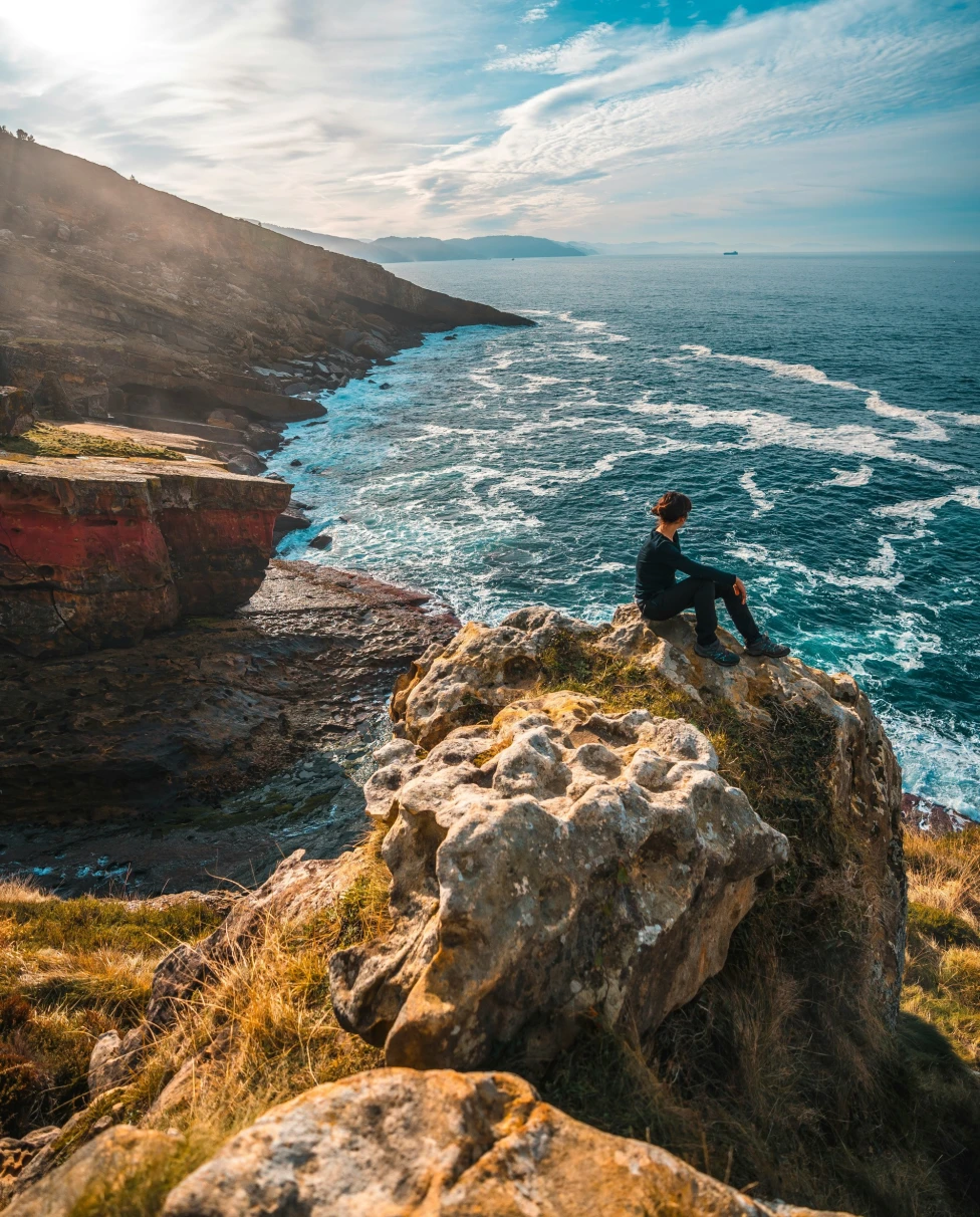 The image depicts an individual sitting on a rocky outcrop overlooking a vibrant blue sea with a clear sky above.