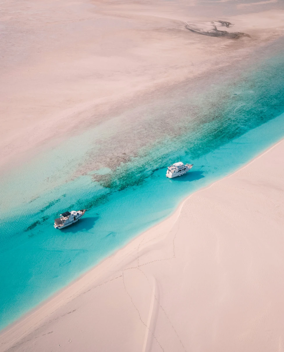 This image depicts an aerial view of two white boats on a body of water surrounded by white sand.
