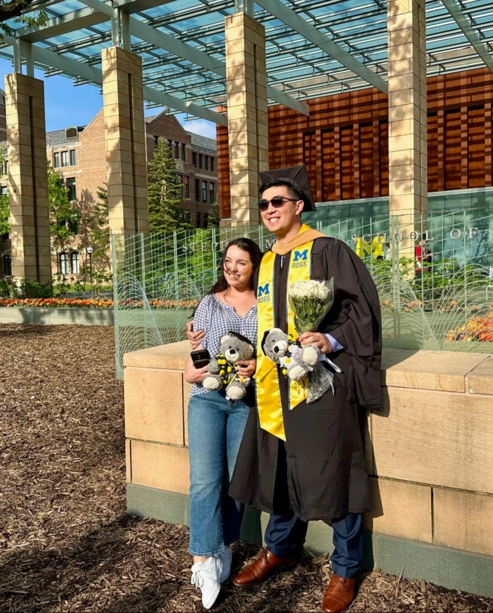 A graduate in cap and gown posing with another person for a photograph outside during the daytime