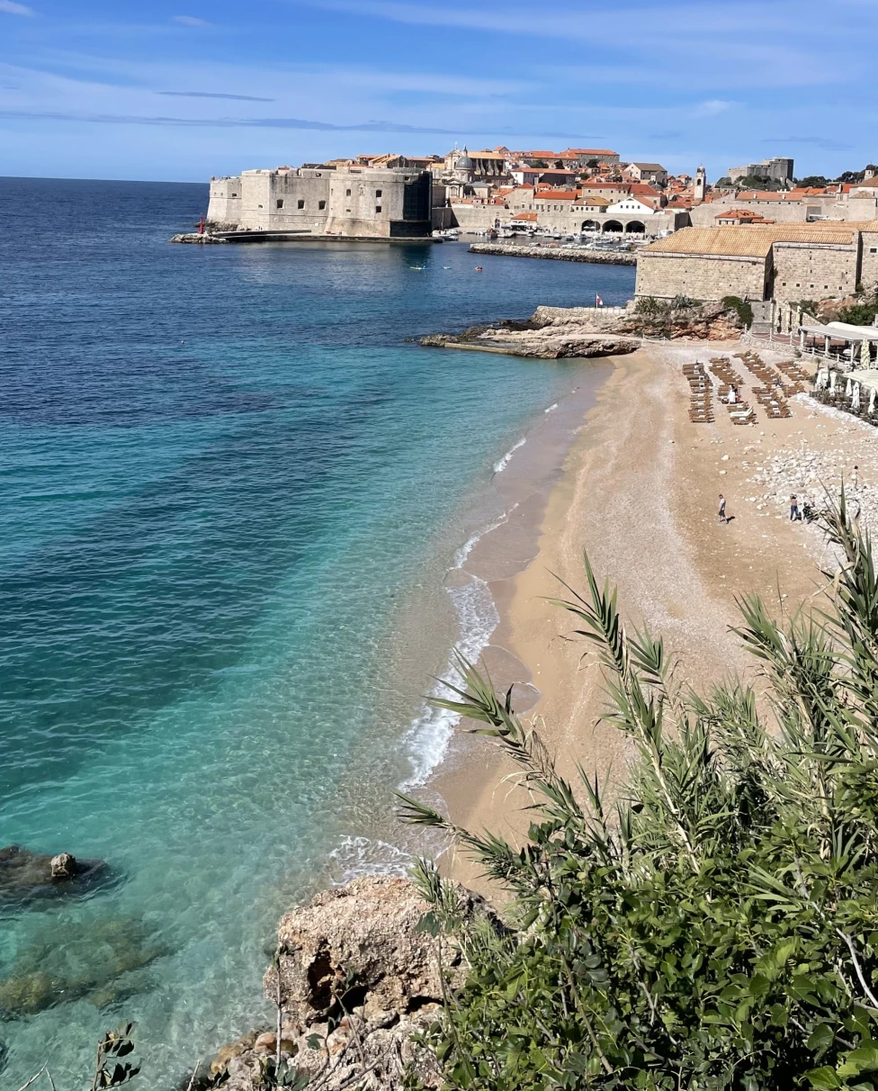 The image captures a picturesque coastal town in Croatia with historic architecture, clear blue waters and a sandy beach under a bright sky.
