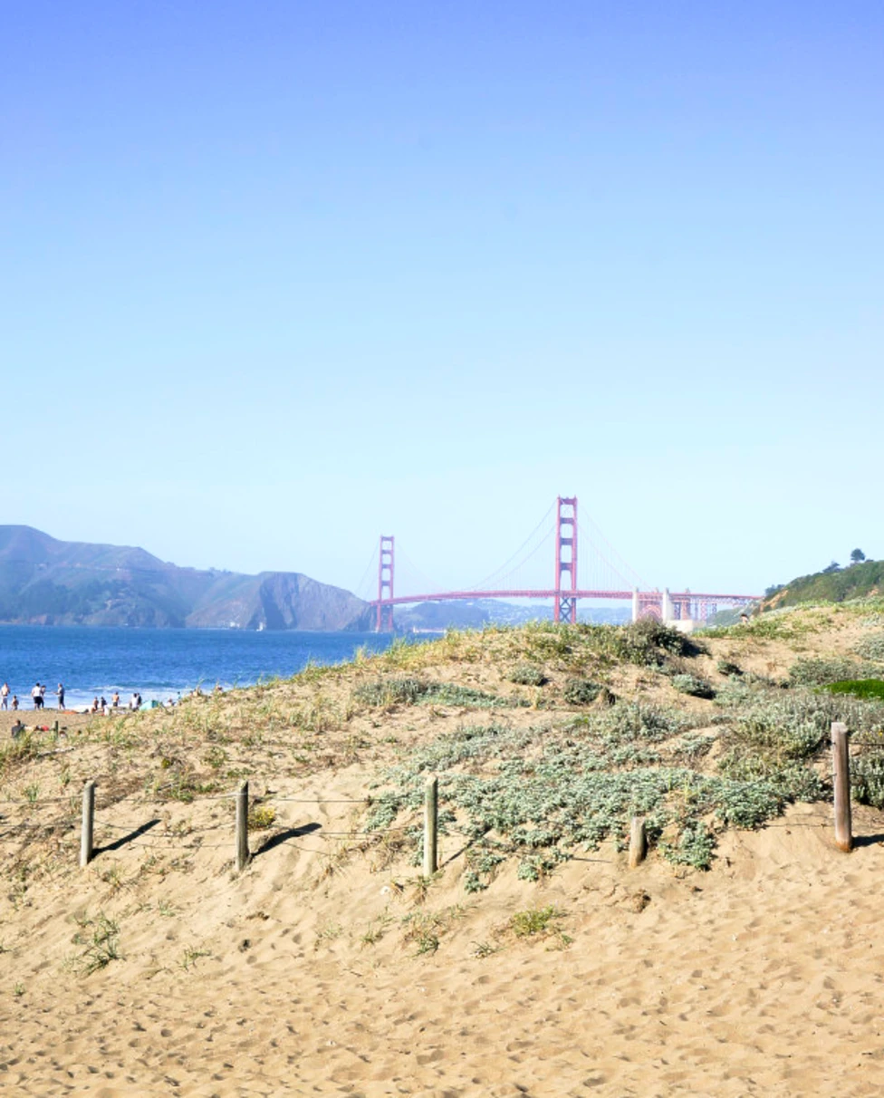 A beach during the daytime with a view of a large, red bridge in the distance