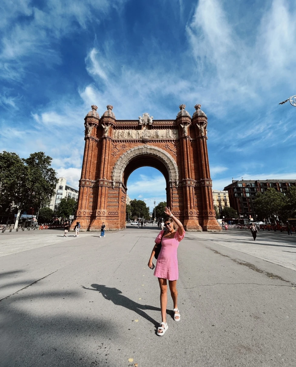A person standing in front of the Arc de Triomf in Barcelona, capturing the grandeur of the monument under a blue sky with scattered clouds.