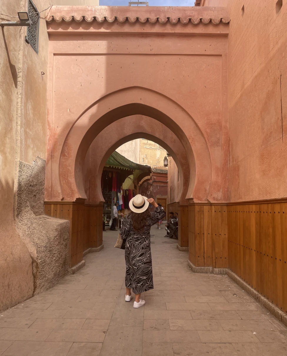 A woman wearing a white hat and a long dress posing under a brown concrete arch during daytime.