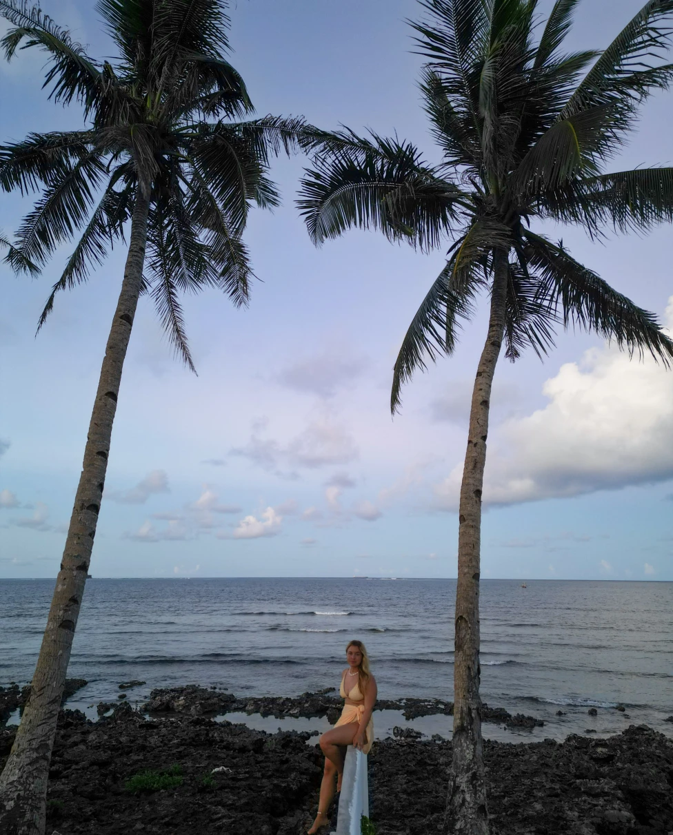 A girl posing near the beach during sunset under two tall palm trees.