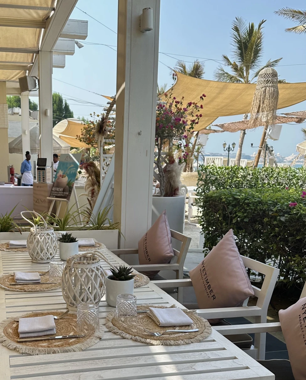 A view of a beautiful outdoor cafe with white tables and chairs that have pink pillows, woven table clothes and white lanterns. There are also sun coverings and palm trees in the background.