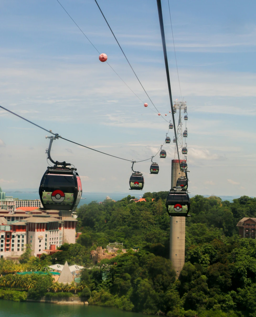 A view of gondolas traveling over the water to Sentosa Island, with trees and Universal Studios in the distance.