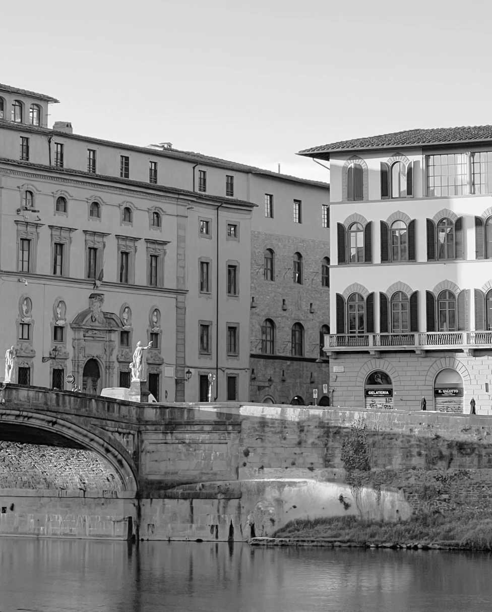 A black-and-white photograph of city buildings in front of a brick bridge over the water