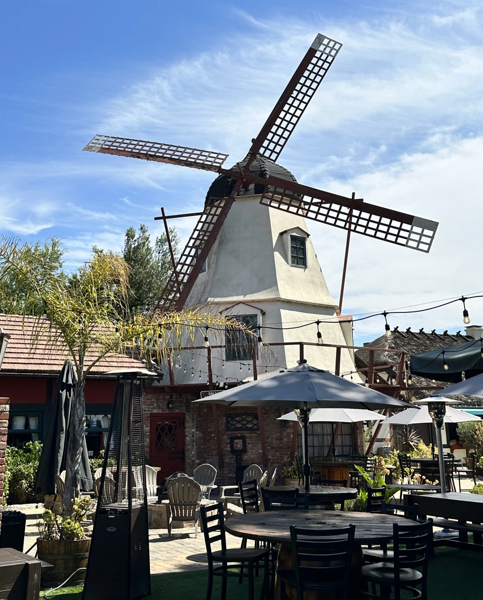 A windmill behind an outdoor patio with umbrellas, tables, chairs and string lights, as well as various plants.