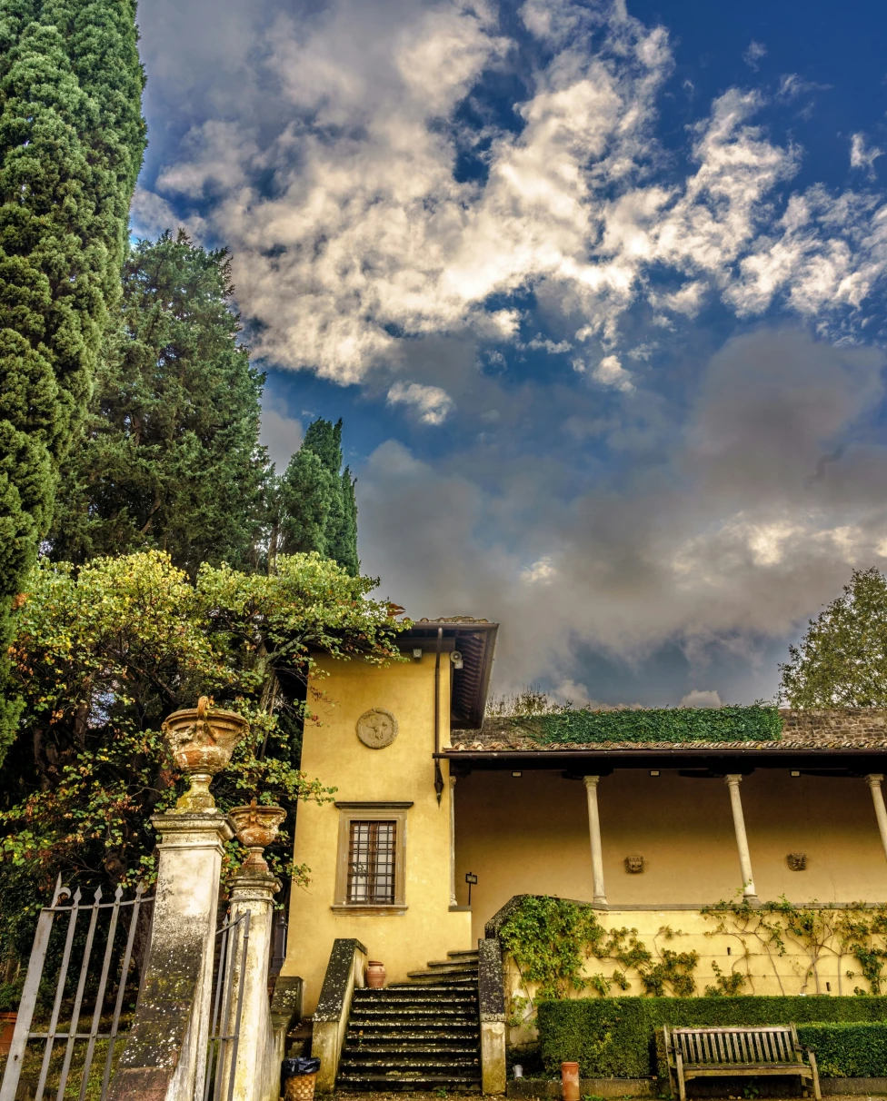 The image features a quaint yellow house with a staircase and lush greenery, under a sky with clouds, exuding old-world charm.