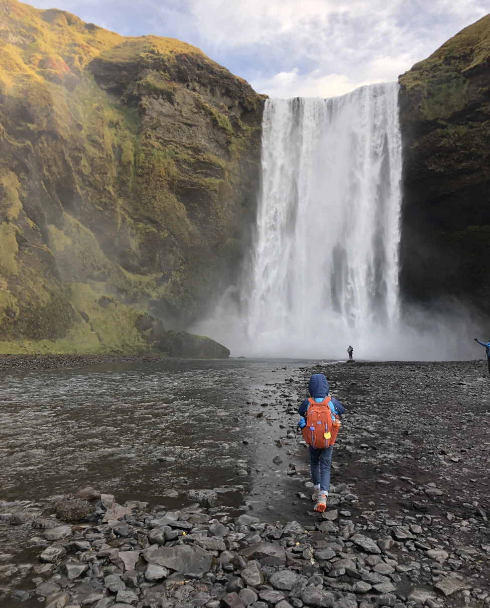 A kid hiking towards Skogafoss Waterfalls on a rocky shore as clouds cover the afternoon sky.