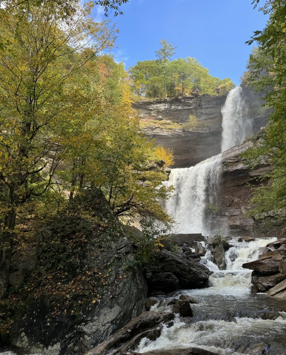 Kaaterskill Falls cascades down a rock face surrounded by lush, fall foliage on a sunny day.