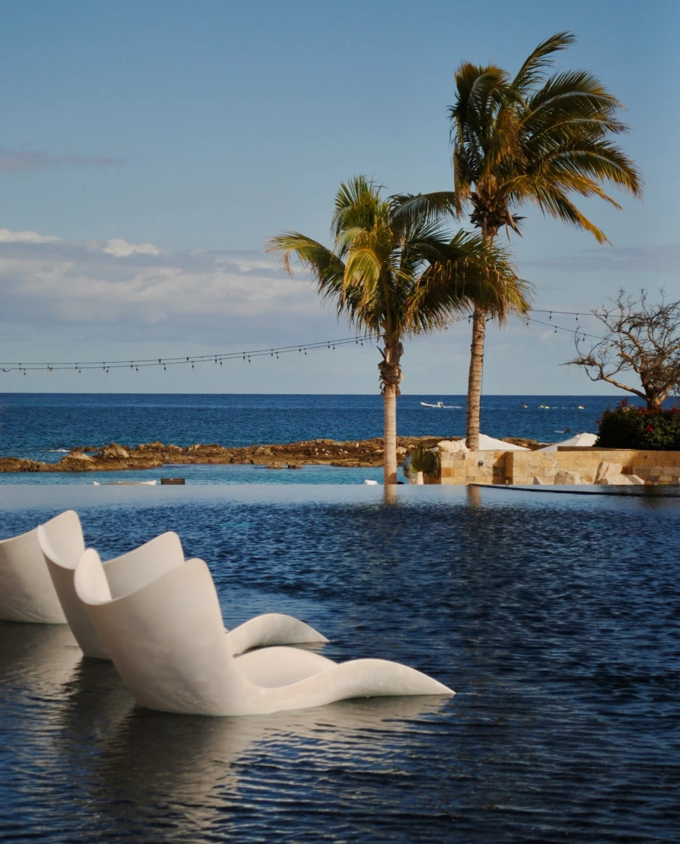 An infinity pool with built-in stone chairs, flanked by palm trees under a clear blue sky and overlooking the ocean, during the best time to go to Cabo.