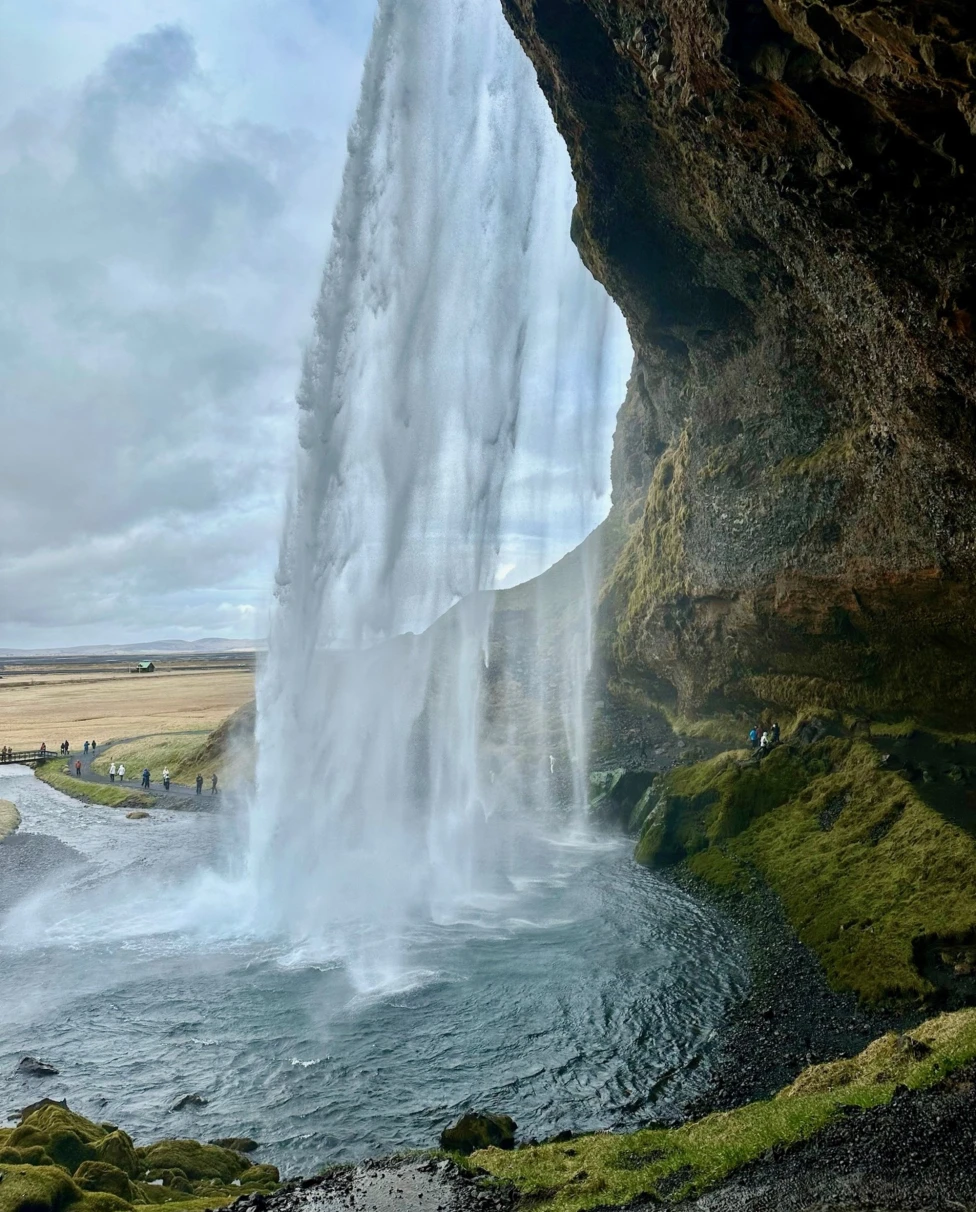 A majestic waterfall cascades down a cliff with a cave visible behind the water curtain, amidst lush greenery.