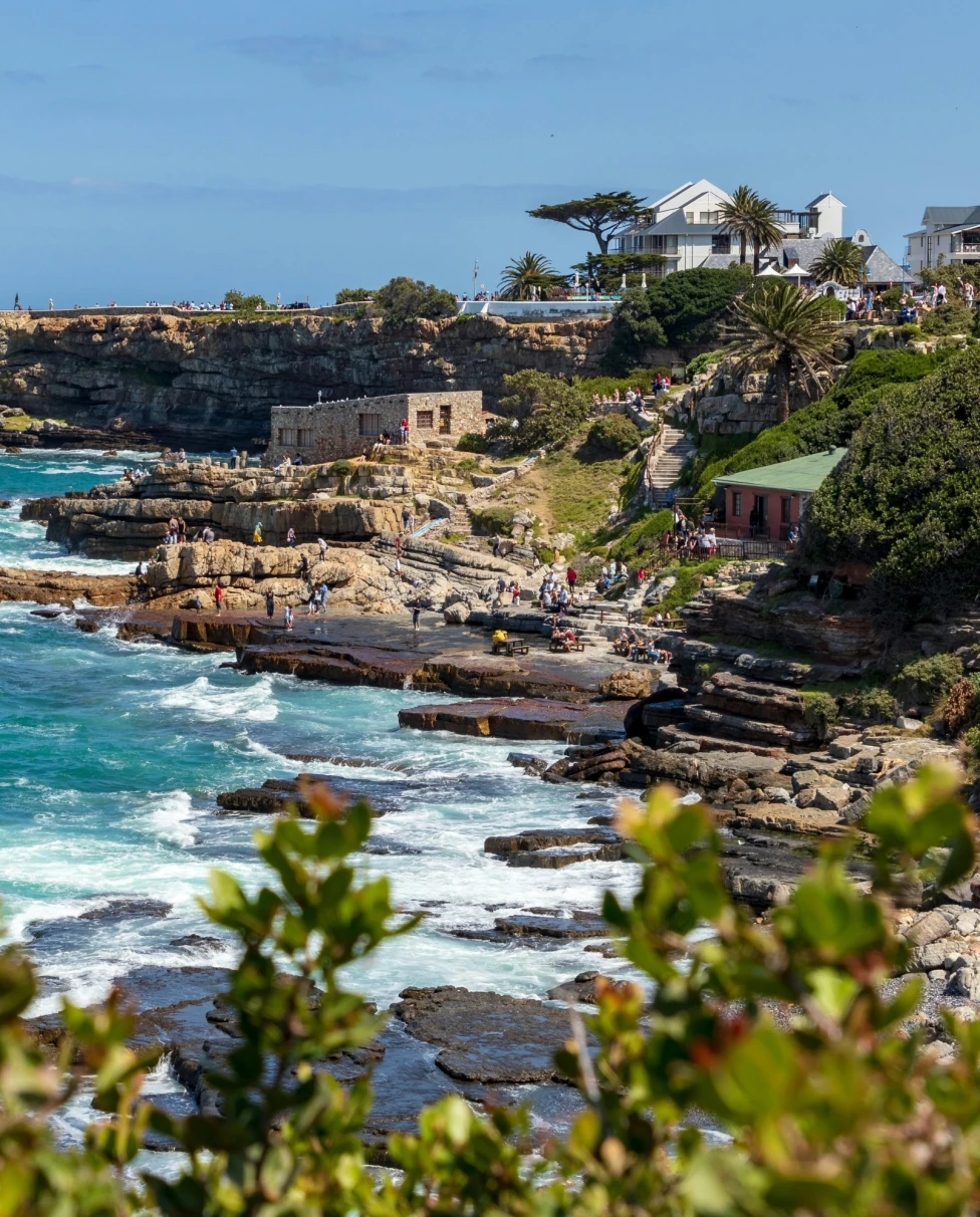 The image depicts a rocky coastline with waves crashing against the shore, vegetation in the foreground, and buildings on the cliff edge under a clear sky.