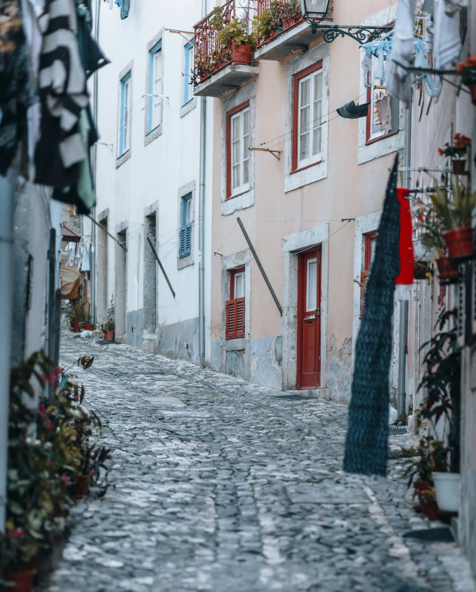 A cobblestone alleyway lined with colorful buildings, greenery and fabric.
