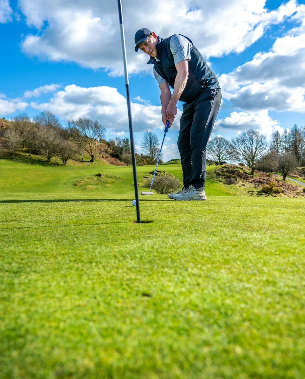 A golfer about to putt on a green, set against a hilly landscape and a partly cloudy sky.