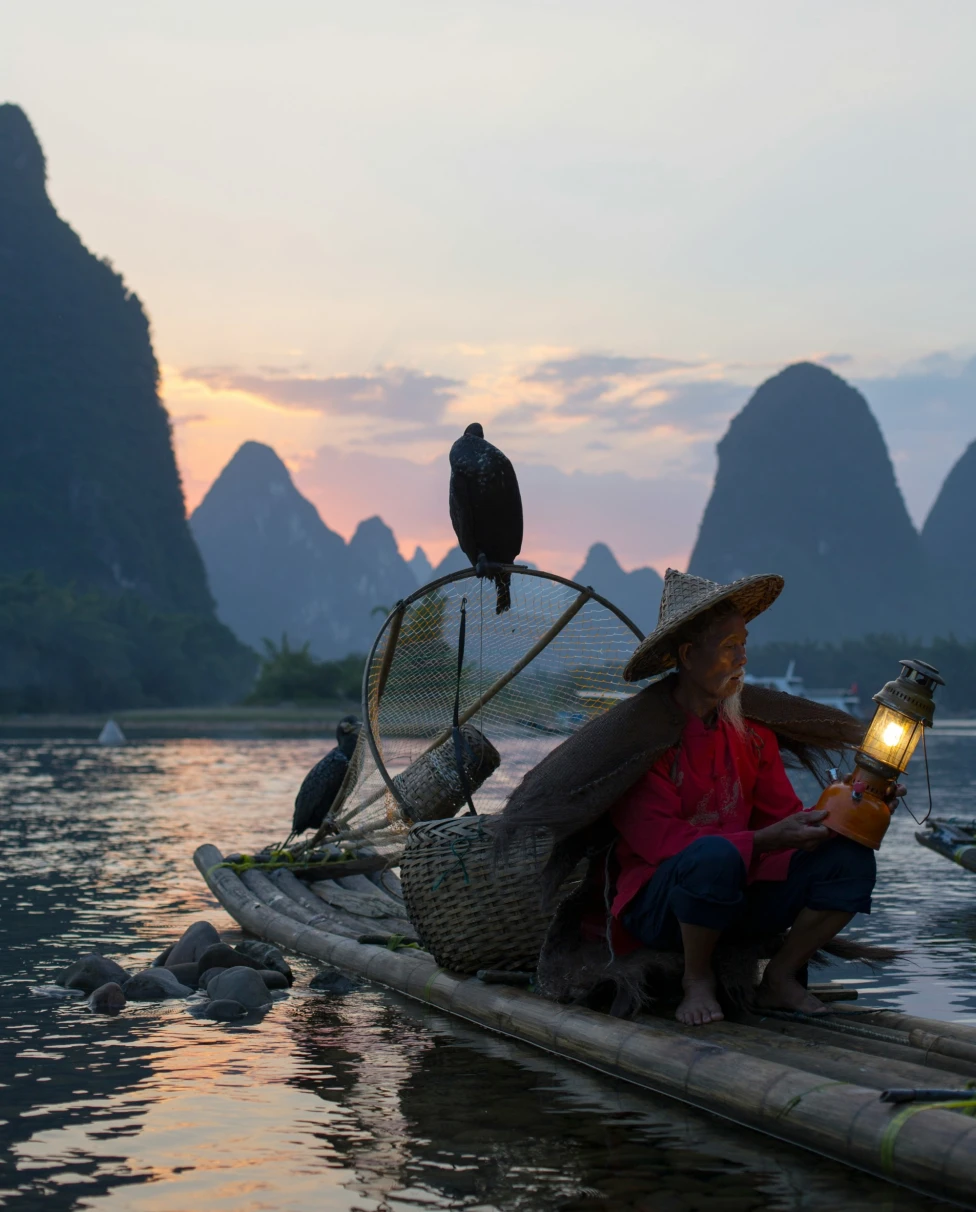 A man sits on a small handmade raft with a lantern at sunset, two birds are perched on the raft with him and there are large rock formations in the ocean ahead of them.