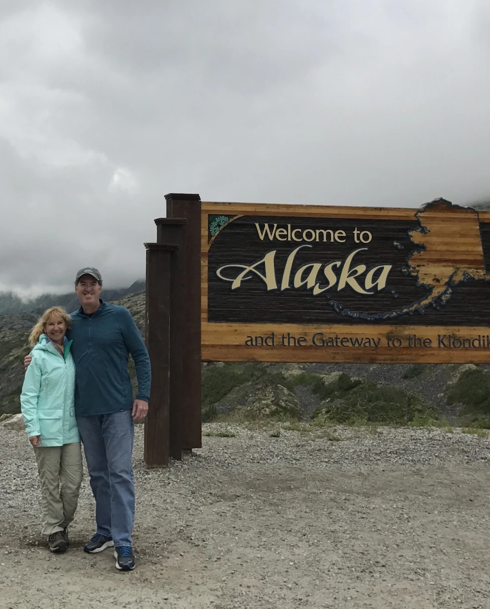 A couple standing in front of a "Welcome to Alaska" sign in front of a mountain landscape, during the best time to travel to Alaska.