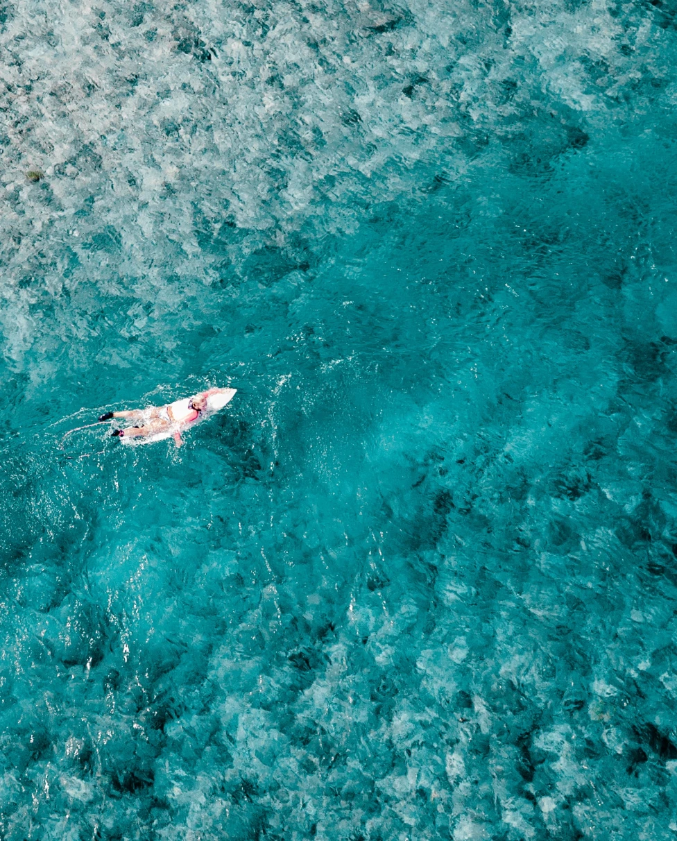 A woman in white on a white surfboard in crystal blue waters with white sand for a luxury surf vacation in the Maldives.