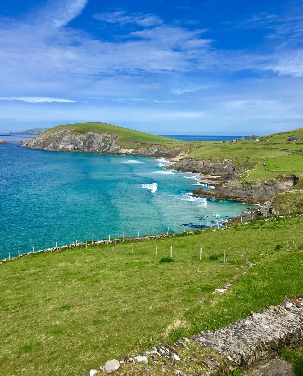 The image shows a scenic coastal landscape with lush green fields, a clear blue sky and a tranquil sea.