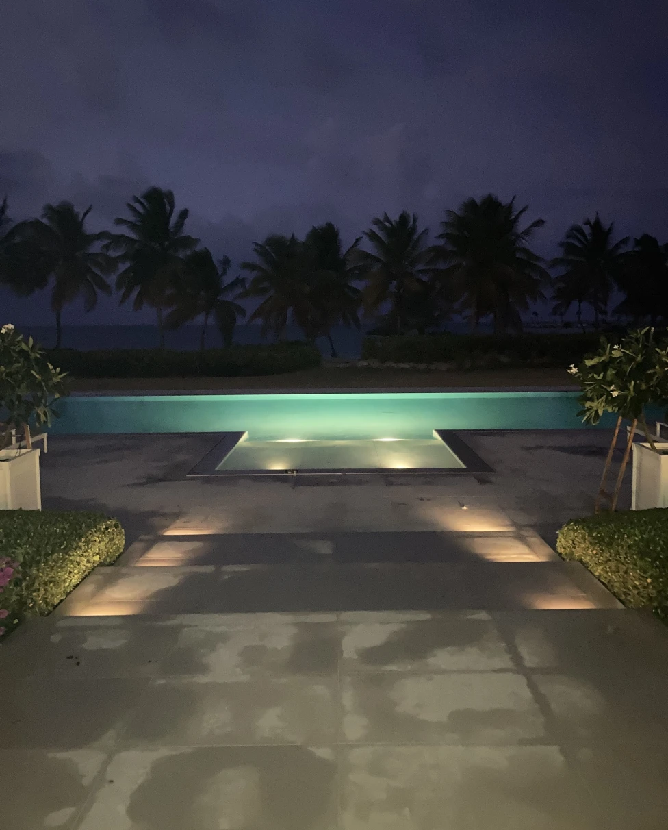 An illuminated swimming pool during twilight with surrounding vegetation and a purple sky.