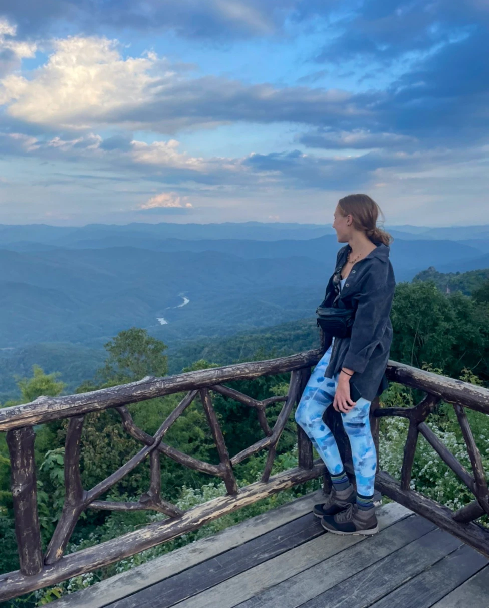 Advisor on a wooden platform overlooking a scenic mountain vista, with a winding river or road below and a partly cloudy sky above.
