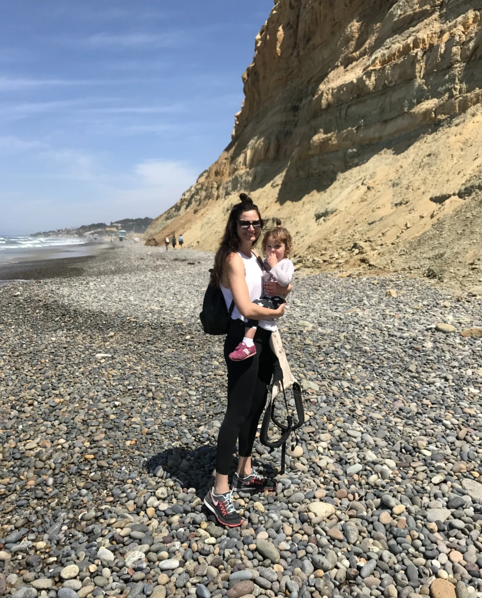 A picture of a woman holding a baby near a beige mountain on top of a rocky beach at daytime.