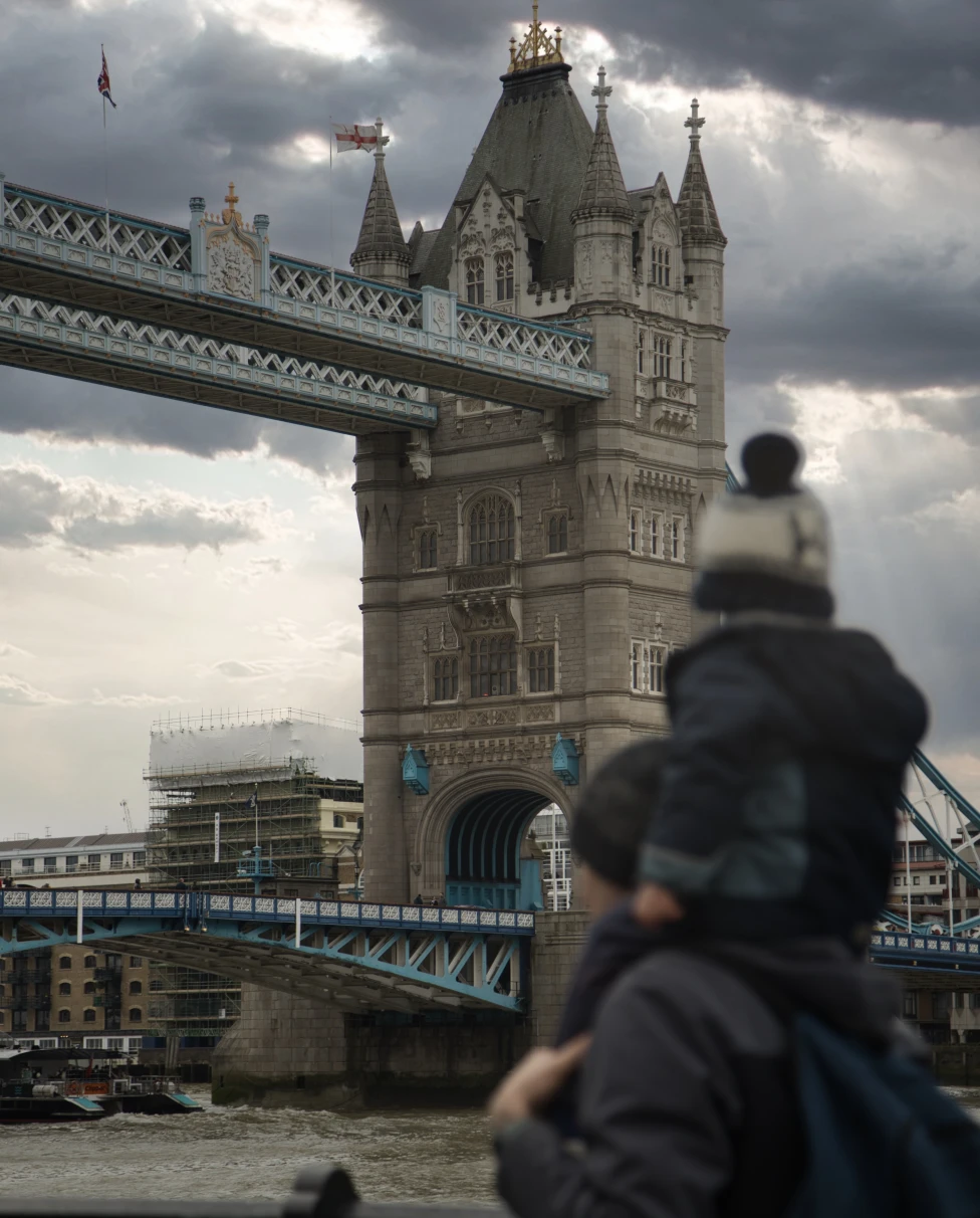 Man with child on shoulders walks past bridge and building on cloudy day