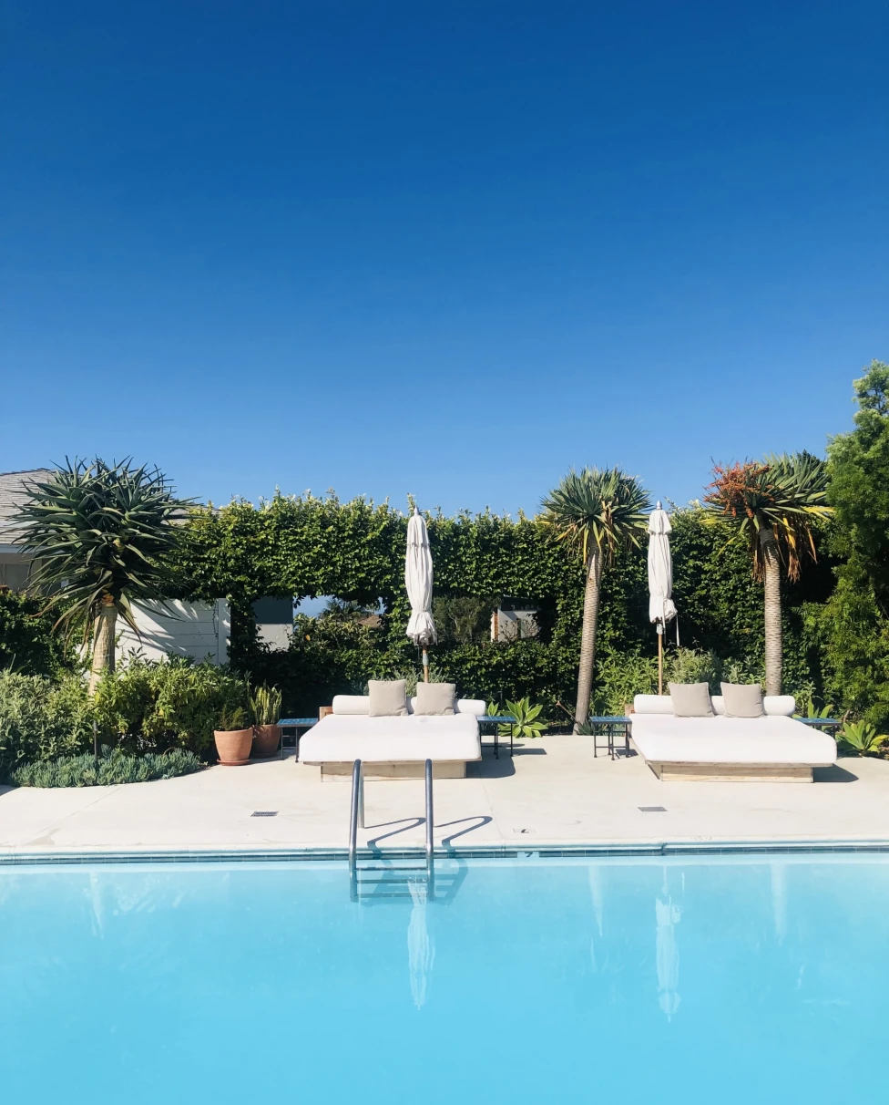 An outdoor swimming pool with sun loungers and a neatly trimmed hedge under a clear blue sky.