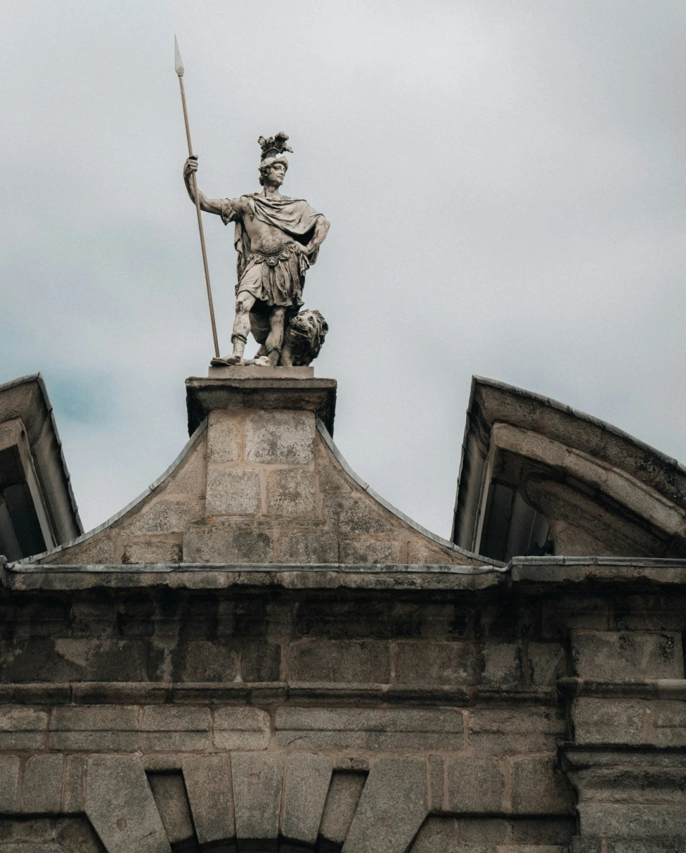The image displays a statue of a warrior with a spear atop an ornate stone pediment against a cloudy sky.
