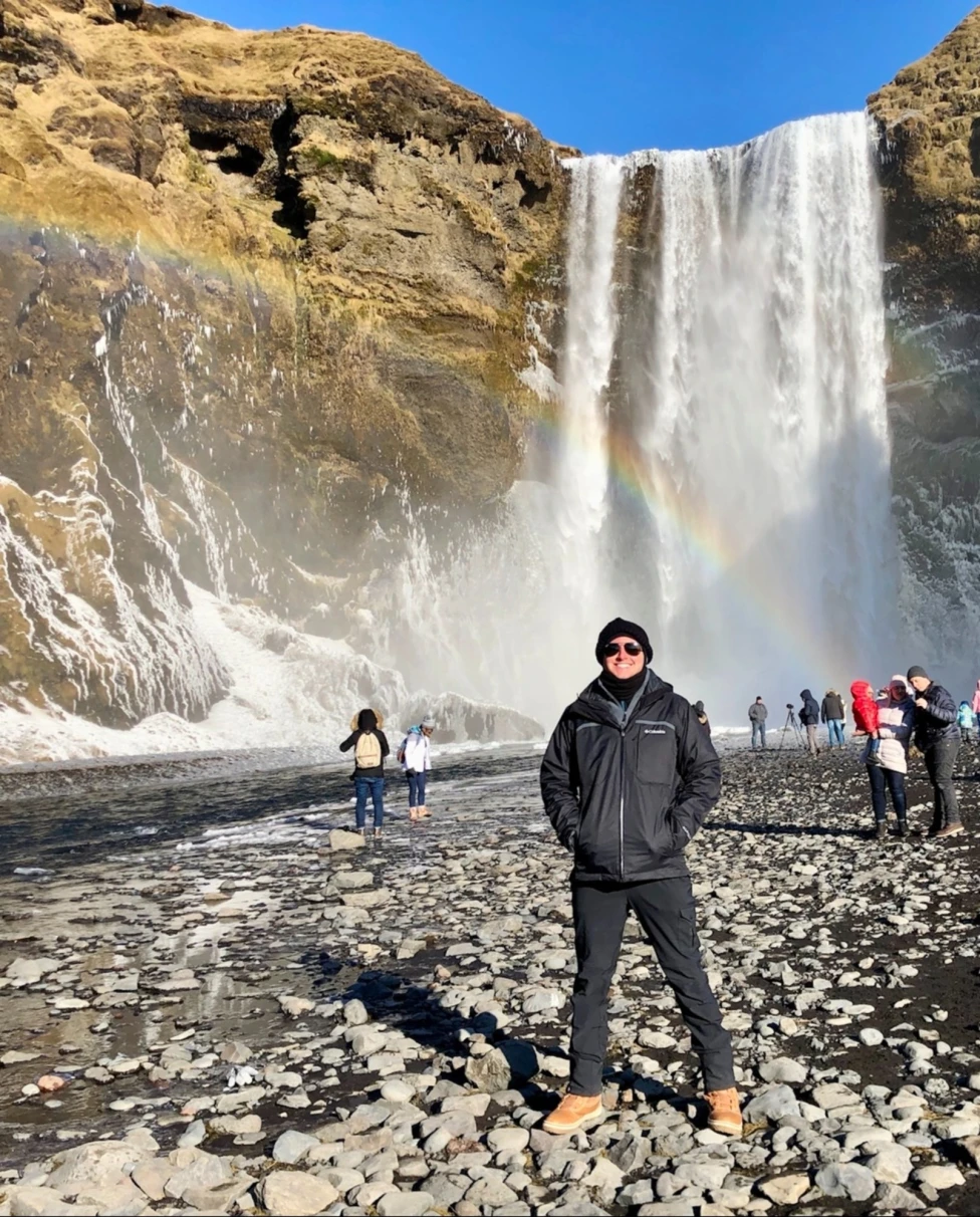The image depicts a person standing in front of a waterfall with a rainbow during the daytime.