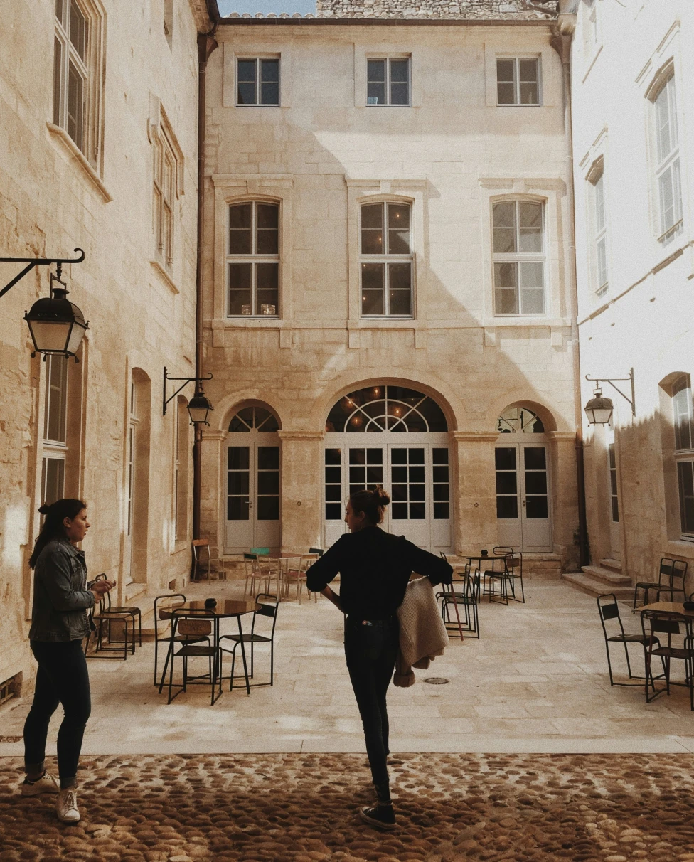 A sunlit courtyard with two people and outdoor seating in front of a classic building.