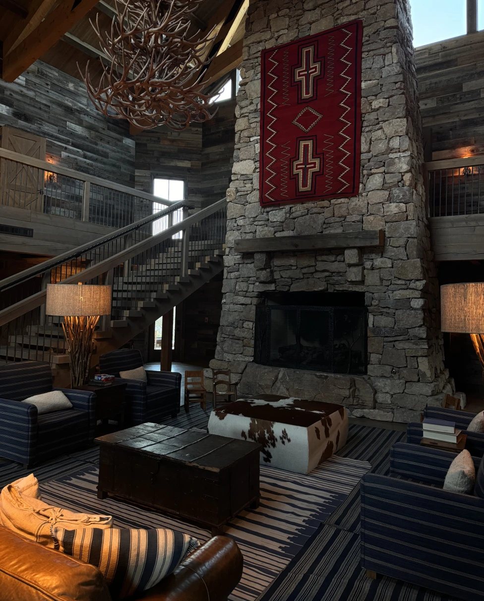 An image of a stone fireplace with a red carpet, wooden chandelier, seating area, leather couch, stairwell and carpet.
