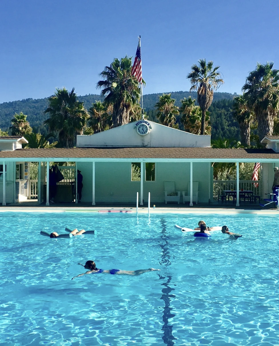 A vibrant blue swimming pool in front of a bright blue building, American flag pole and palm trees. There are people swimming in the pool as well.