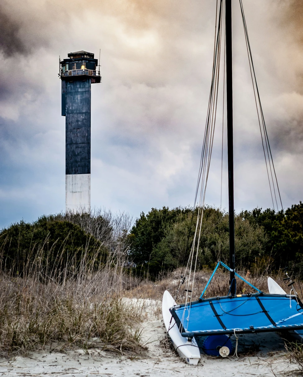 A blue boat on the sand next to a lighthouse with impressive clouds in the background on Sullivan's Island.
