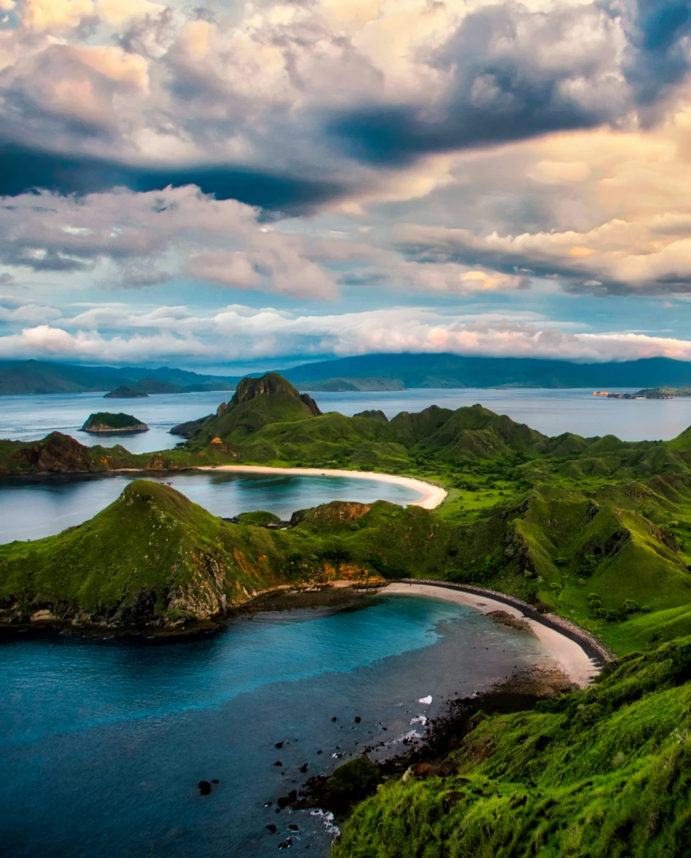 Aerial view of three separate small bays in Komodo National Park with sand in each and green hills surrounding them and clouds ahead