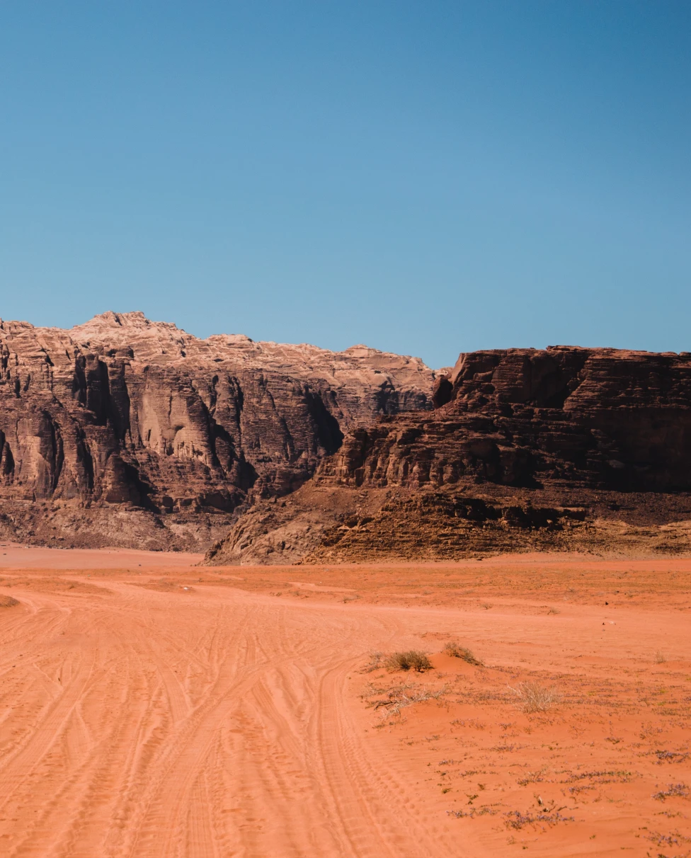 A visit to Jordan with red dirt roads and dark brown and red rock structures against a blue sky.
