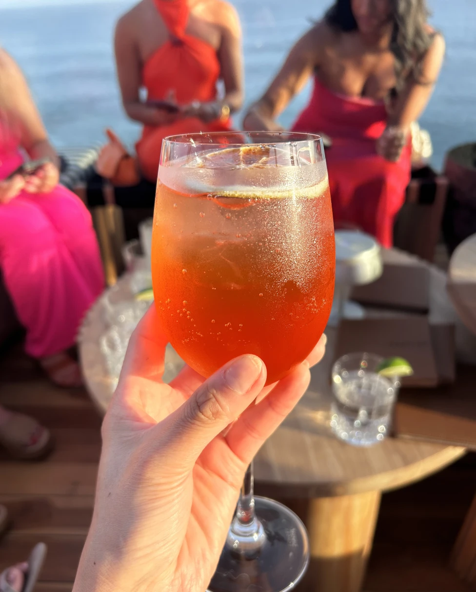 Holding an orange-colored cocktail up at a table with a group of girls in the background.