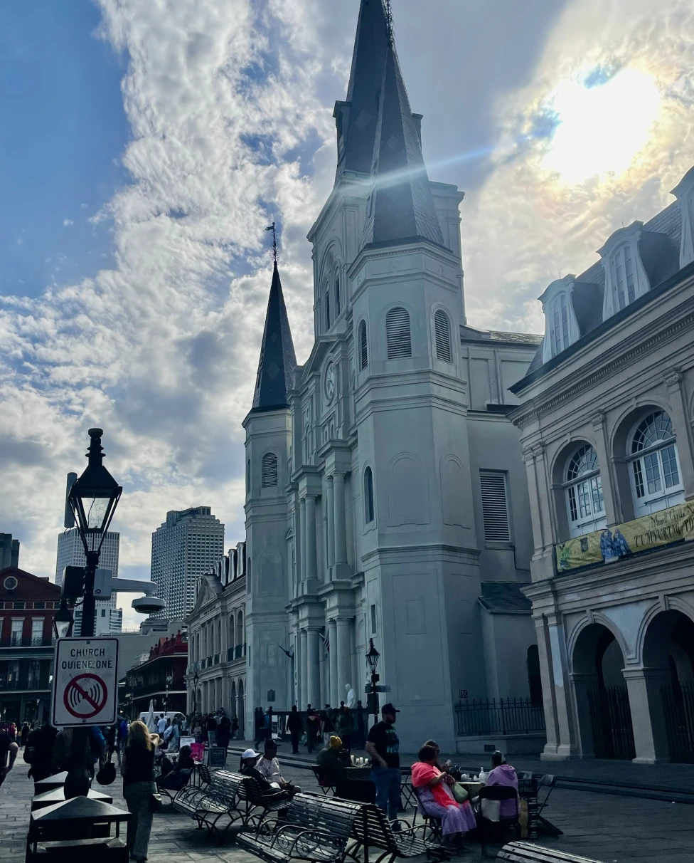 A church with tall steeples and benches in front of it with the sun peeking through clouds behind it.