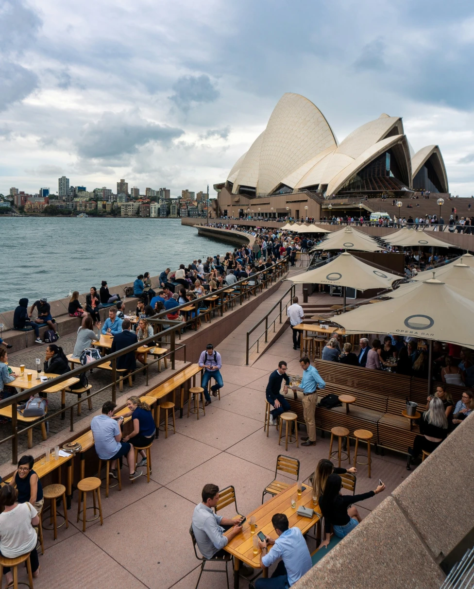 The image depicts an outdoor dining scene with guests enjoying a meal against the backdrop of the Sydney Opera House under a cloudy sky.