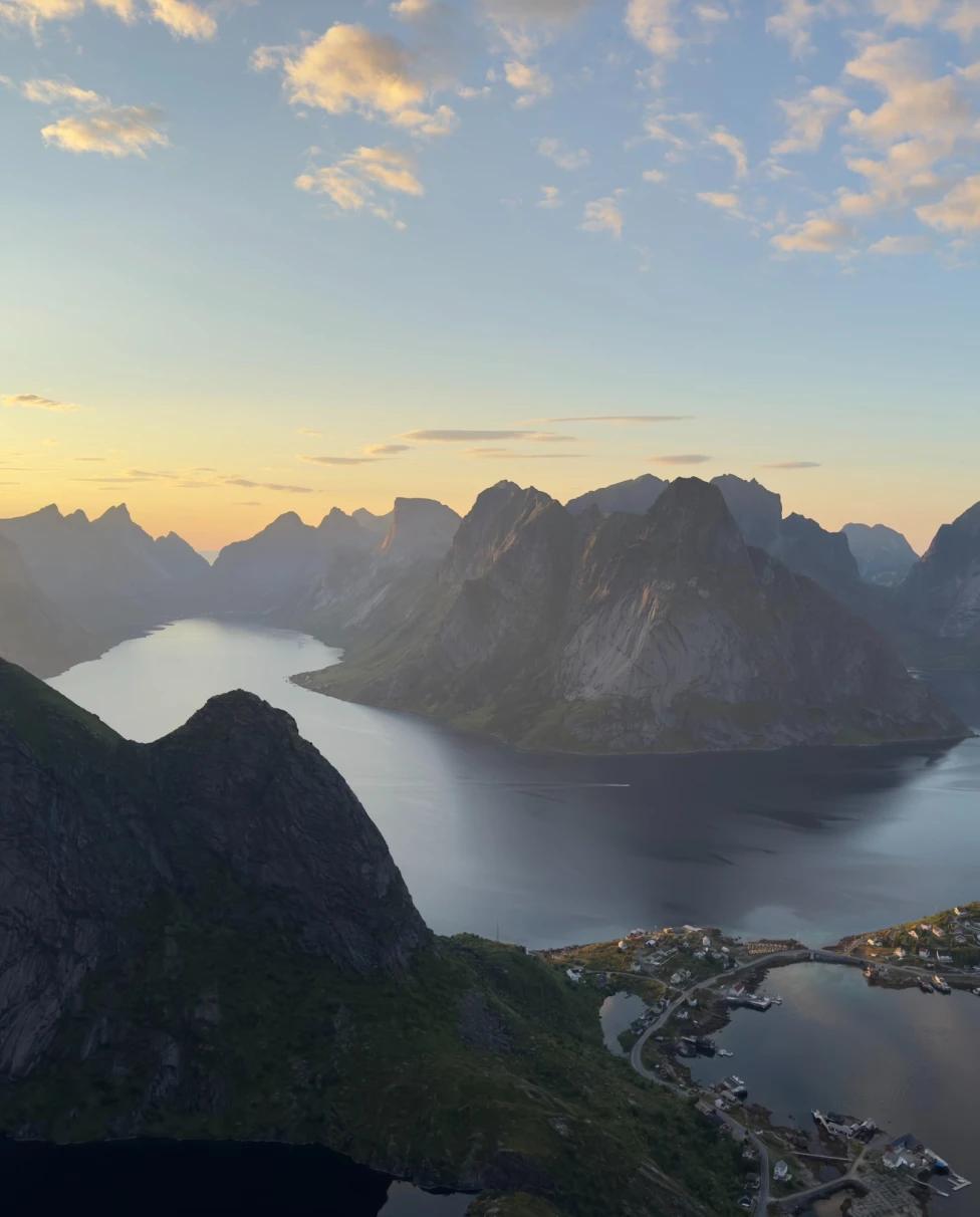 A fjord surrounded by rugged mountains during sunset, with a small settlement at the water’s edge.