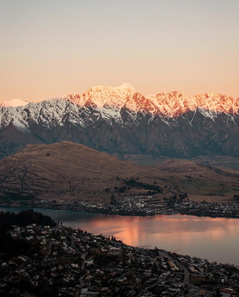 a town on a lake at the base of a snow topped mountain range at sunrise