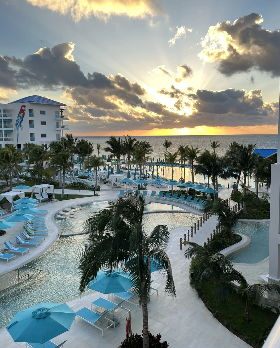 The image shows a sunset view over a resort with a pool and palm trees at sunset.