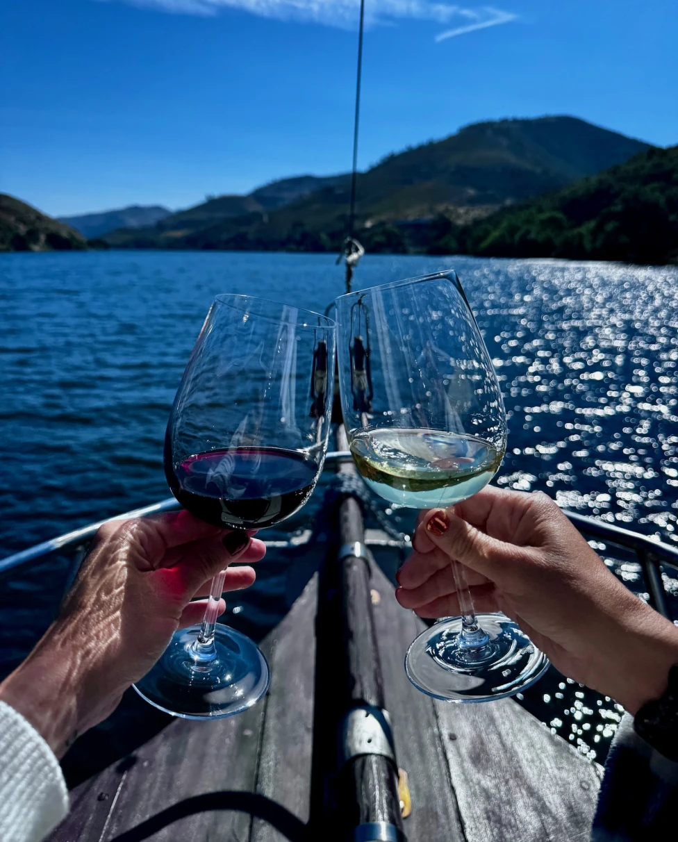 Two people's hands clinking wine glasses while on a boat in a river during the daytime