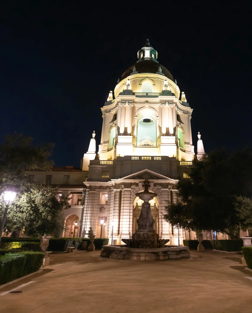 The picture depicts a large white building with a dome, a statue and trees lit up at night time.  It is  Pasadena City Hall.