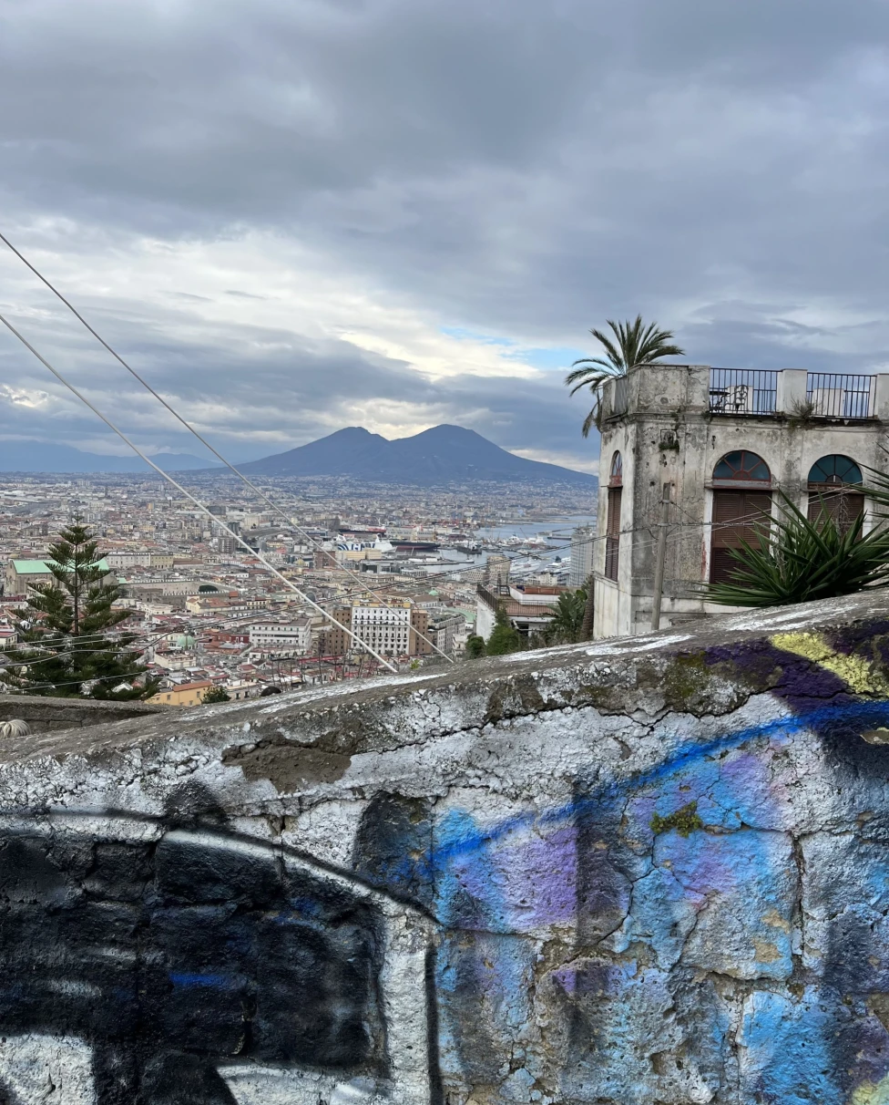 The image shows a panoramic view of a city with Mount Vesuvius in the background, taken from an elevated position with graffiti in the foreground.