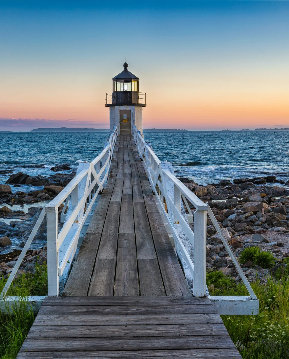 A beautiful wooden bridge with white railings leading to a lighthouse looking over the blue water and light orange sunset.