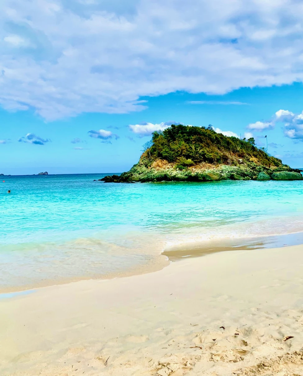 An island dotted with lush foliage sits surrounded by lapping waves just off the beach shore on a sunny day.